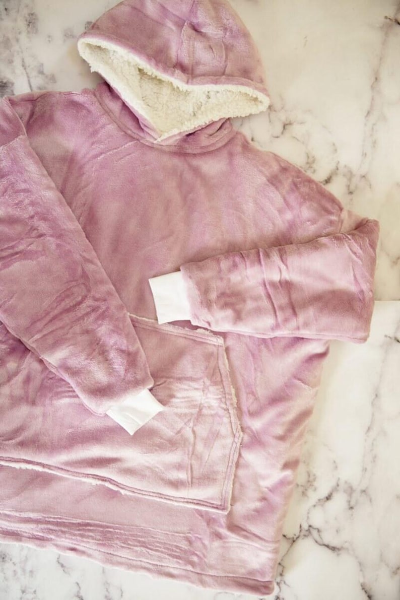 Puede incluir: Una sudadera con capucha de gran tama&ntilde;o hecha de felpa suave en color malva. La prenda cuenta con un forro grueso de sherpa color crema dentro de la capucha y un bolsillo canguro frontal. Tiene mangas largas con pu&ntilde;os blancos acanalados.