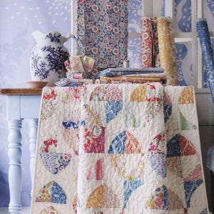 May include: A quilted blanket featuring a repeating pattern of colorful floral fabric semicircles on a cream background, displayed alongside several rolls of matching patterned fabric, a blue and white ceramic pitcher, and folded fabric stacks.