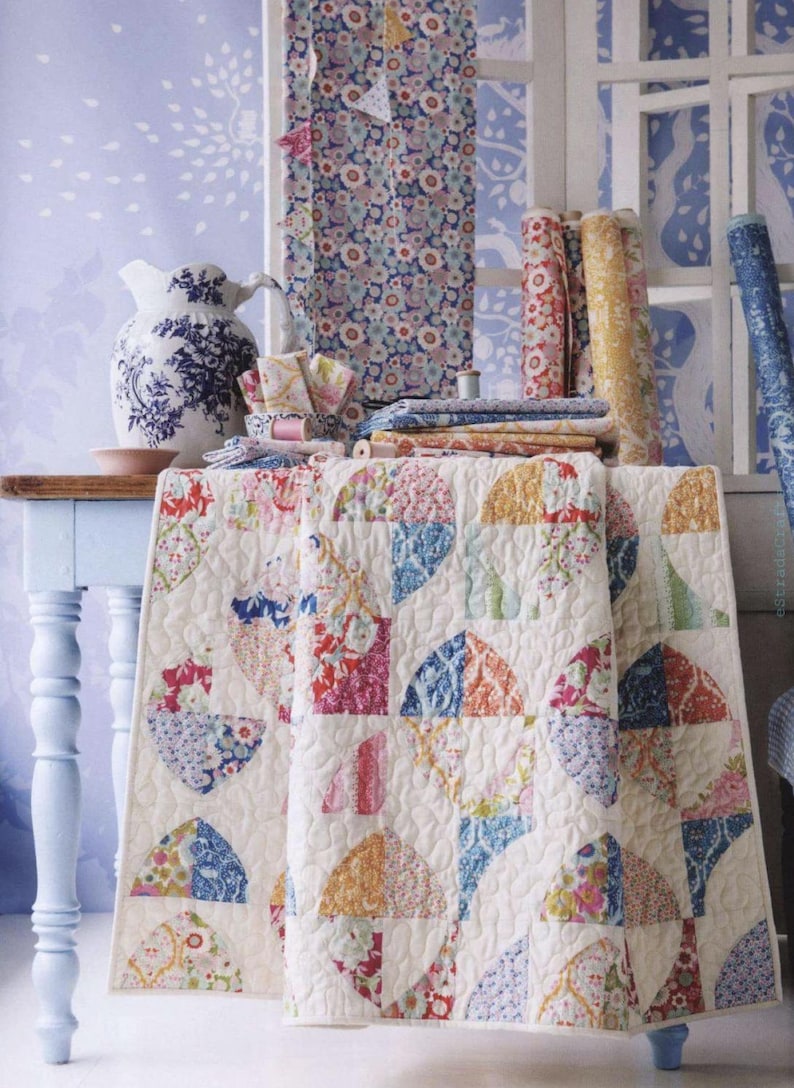 May include: A quilted blanket featuring a repeating pattern of colorful floral fabric semicircles on a cream background, displayed alongside several rolls of matching patterned fabric, a blue and white ceramic pitcher, and folded fabric stacks.