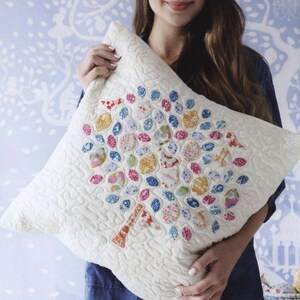 May include: A square off-white quilted throw pillow featuring a central tree applique. The tree trunk is orange and white, while the canopy is composed of numerous small, rounded fabric patches in various floral and geometric patterns in pink, blue, yellow, and red.