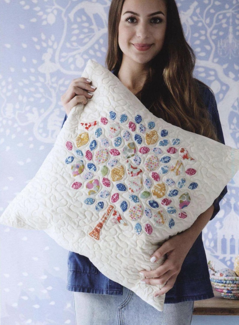 May include: A square off-white quilted throw pillow featuring a central tree applique. The tree trunk is orange and white, while the canopy is composed of numerous small, rounded fabric patches in various floral and geometric patterns in pink, blue, yellow, and red.
