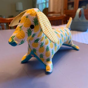 May include: A stuffed dachshund toy made of white fabric with a yellow and green pineapple print. The long ears feature a yellow and white gingham pattern. It has black button eyes, a blue fabric nose, and blue fabric accents on the paws and belly.