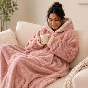 Puede incluir: Una sudadera tipo manta con capucha de gran tama&ntilde;o en color rosa empolvado, hecha de material de piel sint&eacute;tica de felpa con forro de vell&oacute;n sherpa color crema, con mangas largas, un bolsillo frontal grande y capucha, sosteniendo una taza blanca.
