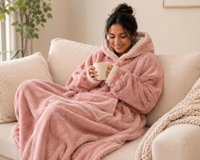 Puede incluir: Una sudadera tipo manta con capucha de gran tama&ntilde;o en color rosa empolvado, hecha de material de piel sint&eacute;tica de felpa con forro de vell&oacute;n sherpa color crema, con mangas largas, un bolsillo frontal grande y capucha, sosteniendo una taza blanca.