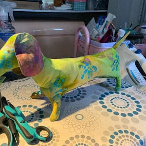 May include: A stuffed dachshund toy made of fabric with a tie-dye pattern in shades of lime green, yellow, and blue. The dog has long, floppy ears with purple and orange accents, a long body, and a short tail, standing on four short legs.