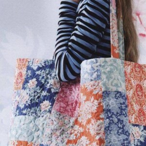 May include: A large quilted tote bag featuring a patchwork design of various floral fabrics in shades of blue, pink, and orange. The bag has two shoulder straps and a rectangular shape with visible stitching patterns across the quilted fabric surface.