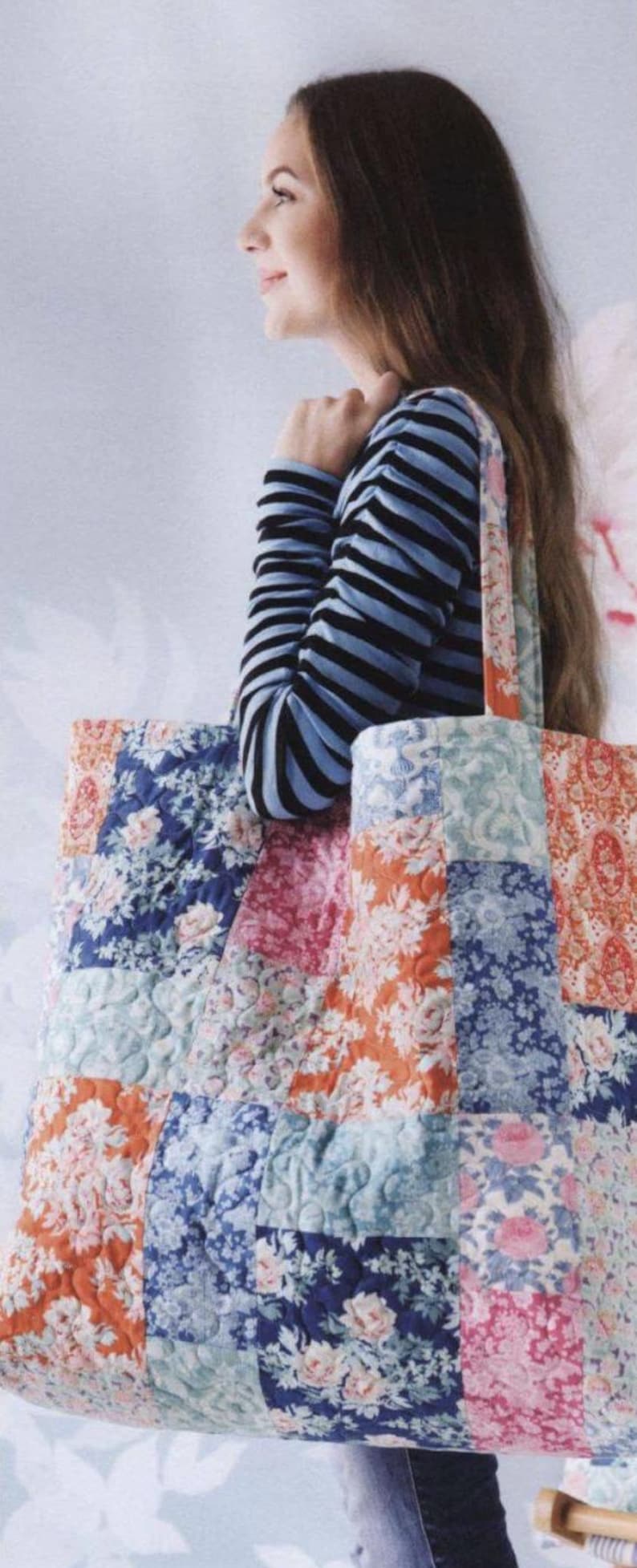 May include: A large quilted tote bag featuring a patchwork design of various floral fabrics in shades of blue, pink, and orange. The bag has two shoulder straps and a rectangular shape with visible stitching patterns across the quilted fabric surface.