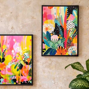 May include: Two framed abstract paintings with colourful floral and geometric designs. The paintings are in black frames and are hanging on a light beige wall.