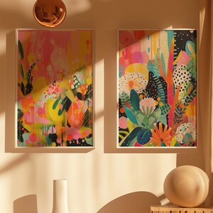 May include: Two framed abstract paintings with colourful floral designs. The paintings are hung on a light beige wall with a white vase and a round, light brown object on a wooden shelf below.