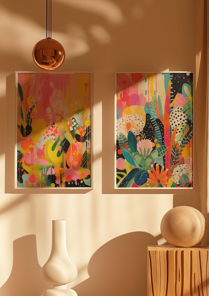 May include: Two framed abstract paintings with colourful floral designs. The paintings are hung on a light beige wall with a white vase and a round, light brown object on a wooden shelf below.