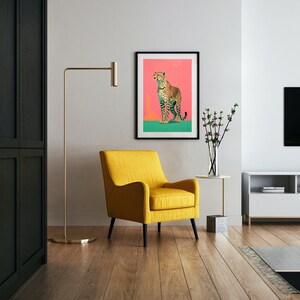 K&ouml;nnte beinhalten: Ein gelber Sessel steht in einem Wohnzimmer mit einem gerahmten Druck eines Geparden auf rosa Hintergrund. Der Druck h&auml;ngt an einer wei&szlig;en Wand. Eine goldene Stehlampe steht neben dem Sessel. Ein kleiner Tisch mit einer Vase mit Blumen steht im Vordergrund.