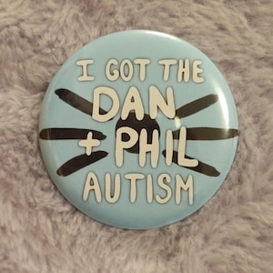May include: A light blue button with the text "I GOT THE DAN + PHIL AUTISM" in white. The button has black lines radiating from the center. The button is round.