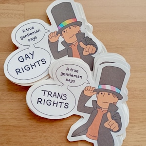 Puede incluir: Un conjunto de pegatinas con un caballero de dibujos animados con un sombrero de copa con una banda arcoíris. Las pegatinas tienen bocadillos que dicen "GAY RIGHTS" y "TRANS RIGHTS". También está presente el texto "A true gentleman says".