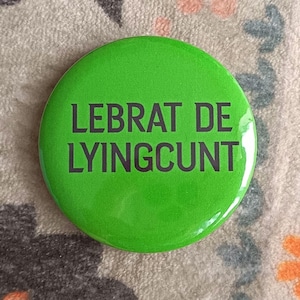 Może przedstawiać: Okrągły przypinany znaczek z jasnym, limonkowym tłem i czarnym tekstem wydrukowanym na środku, który brzmi LEBRAT DE LYINGCUNT. Znaczek ma gładkie, błyszczące wykończenie oraz standardowy okrągły kształt z metalową krawędzią.