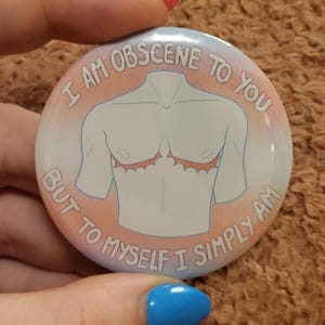 Könnte beinhalten: Ein runder Button mit einem hellblauen, rosa und weißen Design. Der Button zeigt eine stilisierte Illustration eines Torsos mit einer Narbe und dem Text "I AM OBSCENE TO YOU BUT TO MYSELF I SIMPLY AM."