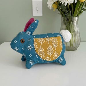 May include: A teal fabric bunny with a white pom-pom tail and a mustard-colored pocket with a white leaf pattern. The bunny has pink inner ears and a brown button eye. It sits on a white surface with flowers in a vase in the background.