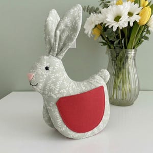 Könnte beinhalten: Ein Stoff-Hase in Hellgrau mit weißem Blattmuster und rotem Fleck. Er hat schwarze Augen und eine rosa Nase. Eine Vase mit Gänseblümchen und Tulpen ist ebenfalls zu sehen.