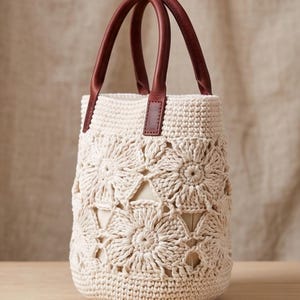 May include: A cream-colored crochet bucket bag featuring a floral motif pattern with openwork details. The bag includes two dark reddish-brown leather handles attached to the top rim, providing a contrast to the light-colored crocheted body.
