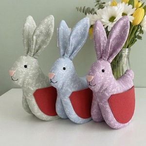 Może przedstawiać: Trzy pluszowe dekoracje w kształcie królików w pastelowych kolorach: zielonym, niebieskim i fioletowym. Każdy królik ma kwiatowy wzór, różowy nos i czerwoną kieszeń. Za królikami stoi wazon z kwiatami.