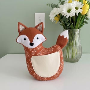 Puede incluir: Un cojín decorativo con forma de zorro en tonos naranja, blanco y beige. El zorro tiene un cuerpo estampado, cara y vientre blancos, y una cola con punta blanca. Las orejas tienen un acento brillante. Un jarrón de flores está en el fondo.
