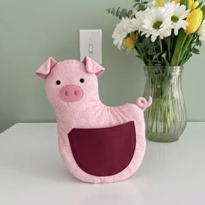 May include: A pink pig-shaped cushion with a burgundy pocket. The pig has a swirled pattern, small black eyes, and pink ears. A vase of white daisies and yellow tulips sits behind the cushion.
