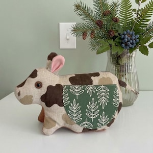 Puede incluir: Un juguete de vaca de peluche con manchas marrones y beige, orejas rosas y un bolsillo verde con un patrón de hojas blancas. La vaca está sobre una superficie blanca.