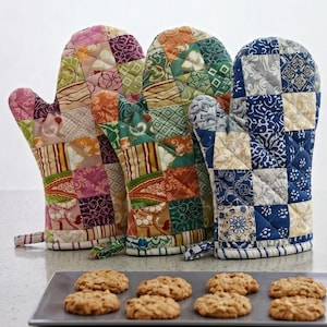 Può includere: Tre guanti da forno trapuntati con motivi patchwork di vari colori, tra cui rosa, verde, arancione e blu, sono in piedi. Una teglia con diversi biscotti dorati è davanti ai guanti, suggerendo un tema di cottura.