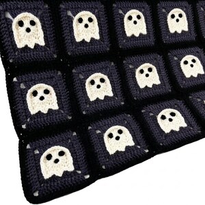 May include: A crochet blanket composed of square motifs, each featuring a cream-colored ghost with two black eyes and a black mouth centered on a dark charcoal background. The squares are joined together to form a repeating grid pattern.