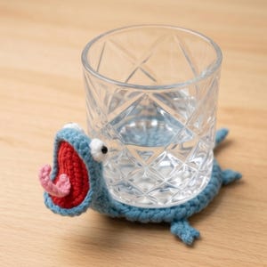 Könnte beinhalten: Ein klares Glas mit Wasser steht auf einem handgefertigten, blauen Häkeluntersetzer in Form eines Cartoon-Monsters. Das Monster hat einen roten Mund, eine rosa Zunge, weiße Augen und blaue Akzente. Der Untersetzer befindet sich auf einer Holzoberfläche.