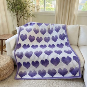 Puede incluir: Una manta de ganchillo blanca y morada con un patrón de corazones. La manta está sobre un sofá blanco. Los corazones tienen un degradado de morado y la manta tiene un borde morado.