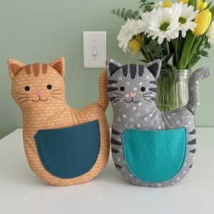 May include: Two stuffed cat-shaped plush toys. One is orange with a subtle pattern and a teal pocket, the other is gray with a white floral pattern, dark gray stripes, and a teal pocket. Both have embroidered black eyes, pink noses, and whiskers.