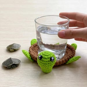 Könnte beinhalten: Ein gehäkelter Schildkröten-Untersetzer in Braun und Grün hält ein klares Wasserglas. Der Untersetzer hat einen Schildkrötenkopf und Flossen. Zwei kleine Schildkrötenfiguren liegen daneben.