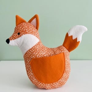 May include: A stuffed fox toy in orange and white. The fox has a patterned body, a white face and chest, and a white-tipped tail. The fox has a black nose and orange ears.