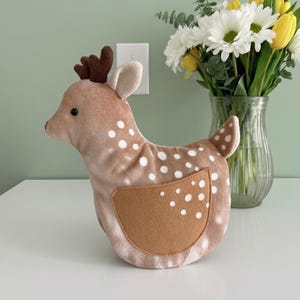 May include: A plush deer-shaped doorstop in shades of tan and brown, with white spots and a brown patch. The deer has small brown antlers and is positioned on a white surface. A vase of white daisies and yellow tulips is in the background.