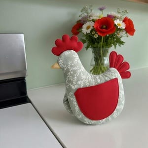 Puede incluir: Un guante de cocina decorativo con forma de gallina. El guante es principalmente gris con un estampado floral blanco, una cresta, una barbilla y una cola rojas, y un parche rojo en la parte delantera. Un ramo de flores está en el fondo.