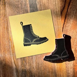 May include: A black boot illustration on a gold card, with a matching boot-shaped cutout. The boot design features laces and a pull loop. The items are placed on a wooden surface.