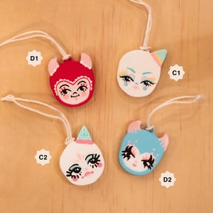 May include: Four whimsical, hand-painted ceramic ornaments with string hangers. Each ornament features a unique face design with colorful details. The ornaments are red, white, and blue, with pink and pastel accents. They are displayed on a light wood surface.
