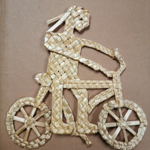 Puede incluir: Una obra de arte de paja tejida que representa a un ciclista en bicicleta. La bicicleta tiene ruedas de radios y manillar. El ciclista tiene el pelo suelto. La obra está hecha de paja natural, sobre un fondo neutro.