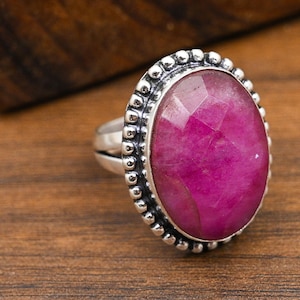 May include: A silver ring featuring a large, faceted oval-shaped ruby gemstone. The ruby is a vibrant magenta color, set in a bezel with a beaded border. The ring is displayed on a wooden surface.
