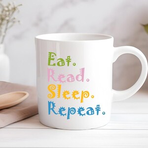 以下が含まれることがあります： カラフルで気まぐれな文字で「Eat. Read. Sleep. Repeat.」と書かれた白いセラミックマグ。マグには湾曲したハンドルがあり、花と木のスプーンのある柔らかい背景に置かれています。