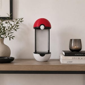Puede incluir: Una l&aacute;mpara cil&iacute;ndrica con un hemisferio superior rojo y un hemisferio inferior blanco, separados por un panel de luz rectangular central. El dise&ntilde;o imita un contenedor esf&eacute;rico con un peque&ntilde;o bot&oacute;n circular blanco en la secci&oacute;n roja frontal.