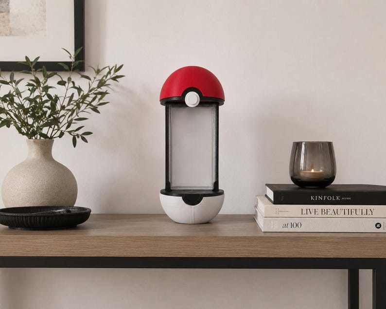 Puede incluir: Una l&aacute;mpara cil&iacute;ndrica con un hemisferio superior rojo y un hemisferio inferior blanco, separados por un panel de luz rectangular central. El dise&ntilde;o imita un contenedor esf&eacute;rico con un peque&ntilde;o bot&oacute;n circular blanco en la secci&oacute;n roja frontal.