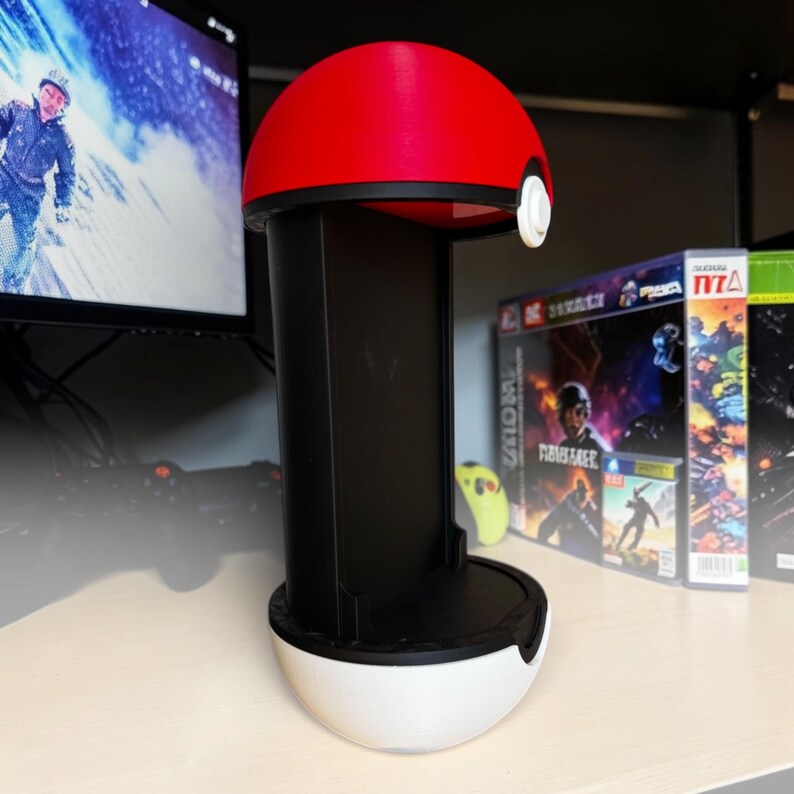 Puede incluir: Un soporte vertical para mando de juego dise&ntilde;ado como una esfera con una mitad superior roja y una mitad inferior blanca. El centro presenta una columna de soporte rectangular negra con un peque&ntilde;o bot&oacute;n circular blanco en la secci&oacute;n roja superior.