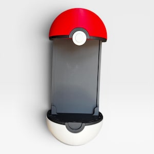 Puede incluir: Un soporte para smartphone dise&ntilde;ado con la forma de una esfera roja y blanca, con una banda horizontal negra y un bot&oacute;n circular blanco en el centro. El dispositivo cuenta con un panel trasero rectangular gris oscuro para sujetar el tel&eacute;fono.