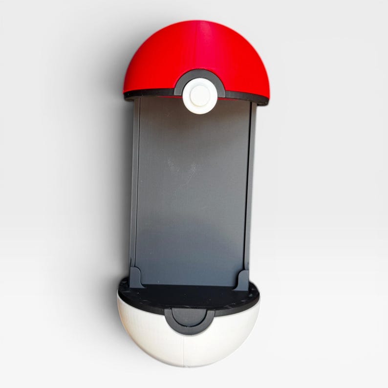 Puede incluir: Un soporte para smartphone dise&ntilde;ado con la forma de una esfera roja y blanca, con una banda horizontal negra y un bot&oacute;n circular blanco en el centro. El dispositivo cuenta con un panel trasero rectangular gris oscuro para sujetar el tel&eacute;fono.