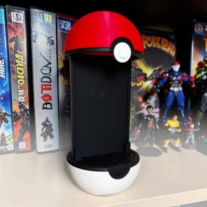 Puede incluir: Un soporte vertical para smartphone dise&ntilde;ado para parecerse a una bola esf&eacute;rica roja y blanca con una banda negra y un bot&oacute;n central blanco. El soporte cuenta con un panel trasero rectangular negro y una base negra para mayor estabilidad.