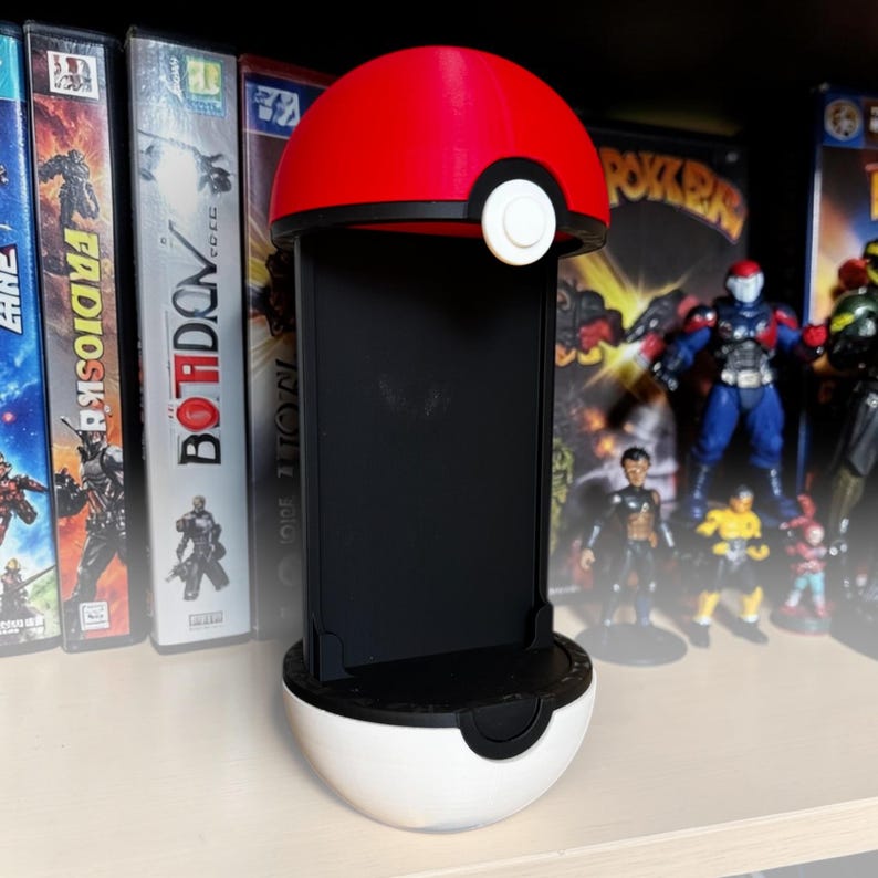 Puede incluir: Un soporte vertical para smartphone dise&ntilde;ado para parecerse a una bola esf&eacute;rica roja y blanca con una banda negra y un bot&oacute;n central blanco. El soporte cuenta con un panel trasero rectangular negro y una base negra para mayor estabilidad.
