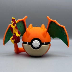 Könnte beinhalten: Eine 3D-gedruckte orangefarbene und hellbraune Charizard-Pokéball. Der Ball hat grüne Flügel, orangefarbene Hörner und eine Flamme. Der Ball ist mit einem weißen Knopf und schwarzen Akzenten versehen.