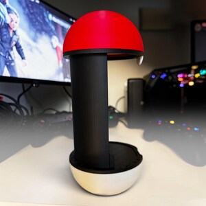 Puede incluir: Un soporte vertical para mando con dise&ntilde;o esf&eacute;rico que presenta una mitad superior roja, un pilar central negro y una base inferior blanca. La estructura tiene forma de bola con una secci&oacute;n central hueca dise&ntilde;ada para sostener un mando.