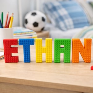 Può includere: Il nome "ETHAN" è composto da lettere colorate a forma di blocchi, disposte su una superficie di legno. Le lettere sono rosse, blu, gialle, verdi e arancioni. Blocchi aggiuntivi di vari colori sono sparsi nelle vicinanze. La scena suggerisce una cameretta.